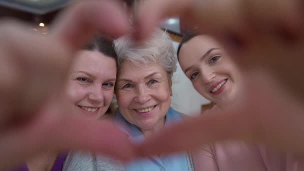 Detailní záběr ženské ruce ve tvaru srdce s úsměvem dospělých seniorů a mladé ženy dívá na fotoaparát s úsměvem. Portrét pozitivní šťastné kavkazské multigenerační rodiny pózující doma. — Stock video