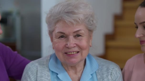 Fejkép egy boldog ősz hajú nőről, aki úgy mosolyog a kamerába, mint a nyugdíjas lánya és unokája. Közelkép az örömteli kaukázusi nyugdíjasról, aki a családjával találkozik. — Stock videók