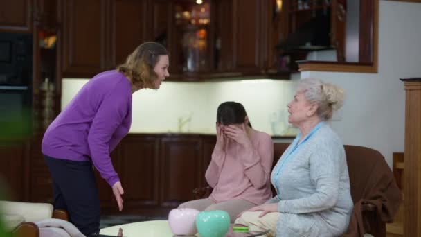 Arg mor och mormor grälar skrikande gestikulerande som upprörd tusenårig kvinna sitter med huvudet i hand. Flergenerationsgräl mellan vita i familjen inomhus. Konfliktbegrepp. — Stockvideo
