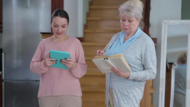 Mulher sênior ler livro como jovem millennial Internet surf em tablet de pé em casa dentro de casa. Família caucasiana positiva desfrutando de hobbies na sala de estar e conversando. Conceito de gerações. — Vídeo de Stock