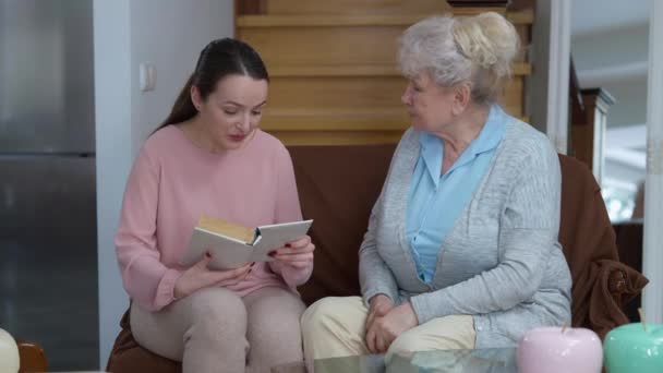 Jonge positieve mooie vrouw lezen boek voor senior dame bespreken verhaal en glimlachen. Portret van een vrolijke ontspannen blanke kleindochter die thuis voor oma zorgt. — Stockvideo