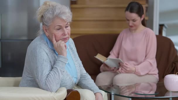 Nachdenkliche ältere kaukasische Frau mit grauen Haaren, die auf einem bequemen Sessel im Wohnzimmer sitzt und der Geschichte lauscht, wie verschwommene junge Millennials im Hintergrund laut ein Buch lesen. Hobby und Generationen. — Stockvideo