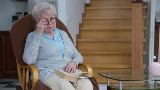 Retrato de una hermosa mujer caucásica de pelo gris leyendo libro quitándose las gafas frotándose los ojos. Relajada anciana jubilada disfrutando del ocio en casa en la sala de estar en el interior. Jubilación y estilo de vida. — Vídeos de Stock