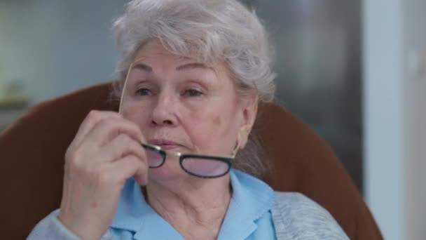 Wajah close-up dari cantik senior Kaukasia perempuan pensiun melepas kacamata menggosok mata. Potret wanita berambut abu-abu duduk di kursi yang nyaman di dalam rumah. — Stok Video