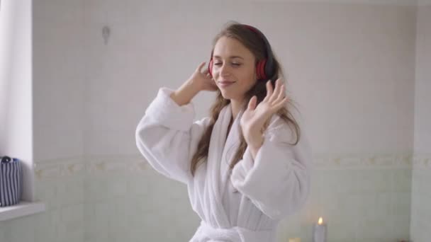 Medium skud af glade unge kaukasiske slank kvinde i badekåbe og hovedtelefoner danser i slowmotion om morgenen. Portræt af glad afslappet munter smuk dame have det sjovt i badeværelset derhjemme. – Stock-video