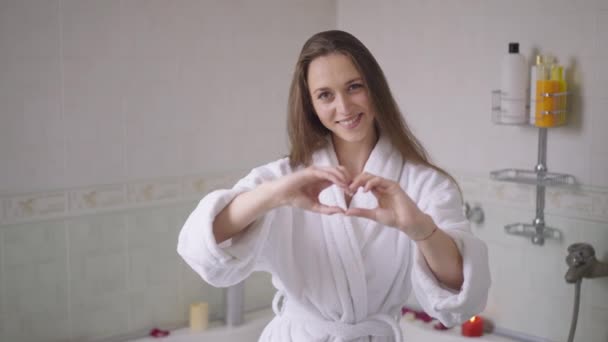 Очаровательная брюнетка стройная молодая женщина, показывающая форму сердца с улыбающимися руками, смотрящими в камеру. Портрет радостной позитивной кавказской дамы тысячелетия, позирующей в замедленной съемке в ванной комнате дома. — стоковое видео
