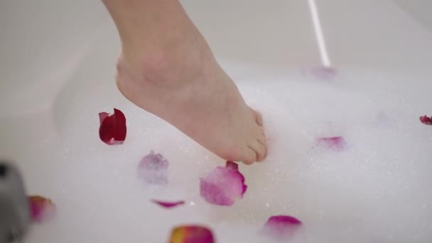 Közelkép vékony női láb megható fürdőkád hab rózsaszirmokkal. Felismerhetetlen fiatal karcsú kaukázusi nő ellenőrzi a víz hőmérsékletét a romantikus fürdőkádban otthon. — Stock videók