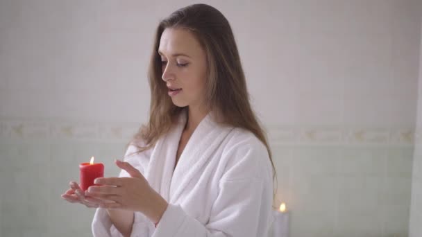 Sensual romântico jovem segurando vela de pé no banheiro em casa sonhando. Retrato de magro bela senhora caucasiana positiva em roupão branco desfrutando de lazer dentro de casa. Movimento lento. — Vídeo de Stock