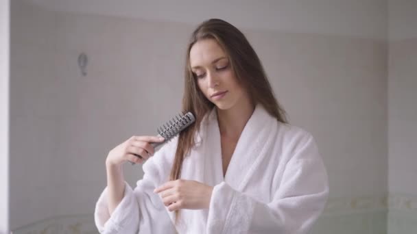 Joven morena caucásica sonriente peinando el pelo en cámara lenta de pie en casa en el baño. Retrato de mujer delgada y segura de sí misma disfrutando de la rutina en el interior de la mañana. Estilo de vida y belleza. — Vídeo de stock