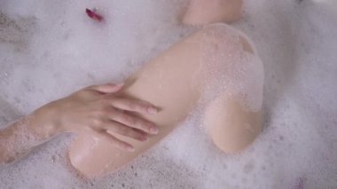 Yakın plan kadın eli, banyo köpüğüyle yavaş çekimde ince bacağı okşuyor. Tanımlanamayan, beyaz tenli genç bir kadın evde banyo yapıyor. İçeride dinlenen rahat bayanın üst görüntüsü..
