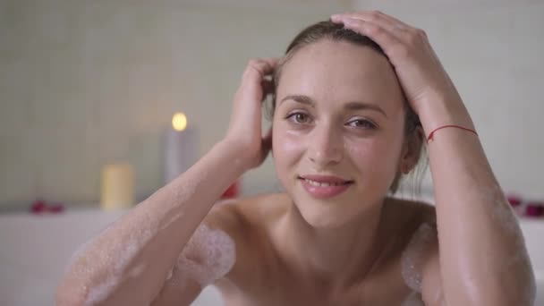 Portret van slanke charmante verleidelijke blanke vrouw die in bad zit en glimlachend naar de camera kijkt. Vooraanzicht van zelfverzekerde sensuele prachtige duizendjarige dame poseren in de badkamer thuis in slow motion. — Stockvideo