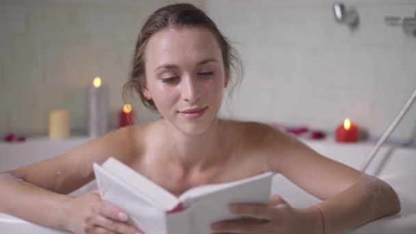 Retrato de la joven caucásica absorbida mujer delgada leyendo libro en la bañera en casa. Encantadora hermosa dama milenaria disfrutando de hobby tomar un baño con velas ardiendo en el fondo. Romance y ocio. — Vídeos de Stock