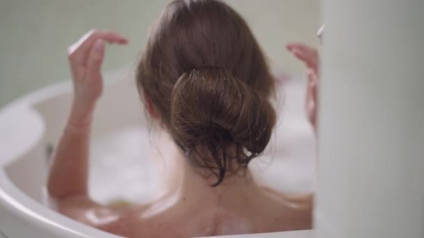 Jeune femme brune touchant les cheveux avec des mains minces en mousse de bain. Vue arrière de dame millénaire caucasienne insouciante détendue profitant de la détente dans la baignoire au ralenti. Concept de loisirs et d'hygiène. — Video