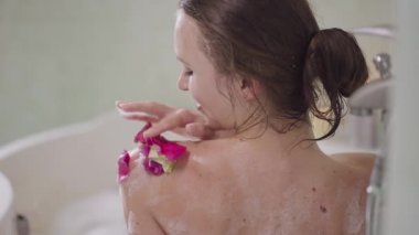 Gülümseyen, sıska, kırmızı çiçek yapraklarını banyodaki köpüğe koyan genç bir kadın. Beyaz tenli romantik kadın evde ağır çekimde banyo yapıyor. Yaşam tarzı ve kadınlık kavramı.