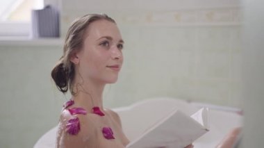 Banyo köpüğü ve kırmızı çiçek yaprakları içinde banyoda kitap okumaktan zevk alan sıska, gülümseyen genç bir kadının yan görünümü. Mutlu, rahat, kaygısız, bin yıllık beyaz kadın içeride banyo yapıyor..