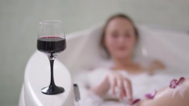 Küvette kırmızı şarap. Arka planda banyo köpüğünün içinde yatan gevşemiş gülümseyen, sıska bir kadın. Kafkasyalı mutlu bayan hafta sonu evde alkolle banyo yapıyor. Yavaş çekim.