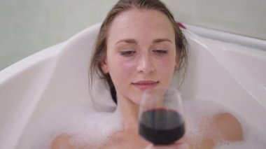 Mutlu, rahat, beyaz, zayıf bir kadının yakından çekilmiş portresi. Ağır çekimde kırmızı şarap içen, köpüklü bir şekilde banyoda yatan. Güzel milenyum hanımefendisi hafta sonu evde, banyoda içki içiyor..