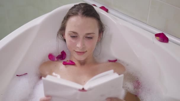 Hoge hoek uitzicht portret van charmante slanke zelfverzekerde vrouw liggend in bad met schuim leesboek glimlachen. Mooie blanke dromerige dame genieten hobby in badkamer thuis in slow motion. — Stockvideo