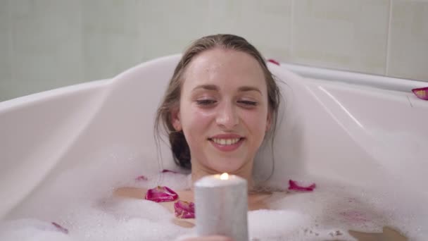 Porträtt av avslappnad leende vacker kvinna liggande i vatten i badkar blåser ut ljus i slow motion. Glad vit smal tusenårig dam njuter av att bada hemma på helgen. Skönhet i hygien. — Stockvideo