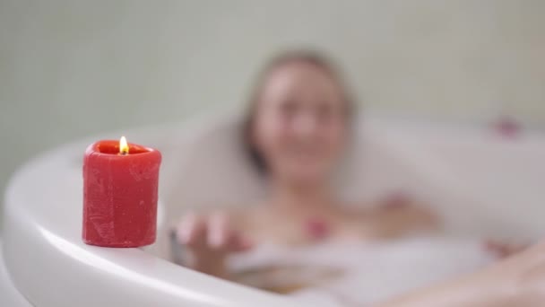 Detailní červená hořící svíčka na vaně s rozmazanou bílou mladou štíhlou ženou koupající se v pozadí. Neznámá dáma si užívá víkendové hygieny ve zpomaleném filmu. — Stock video