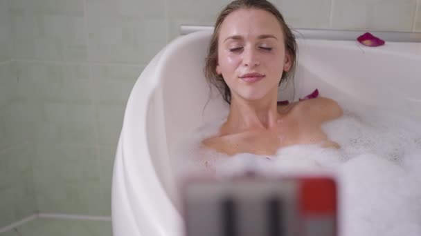Alegre confiante relaxado millennial blogueiro feminino fluindo do banheiro deitado em espuma de banho em beijos de água. Retrato de mulher magra caucasiana despreocupada se divertindo blogando do banheiro em casa. — Vídeo de Stock