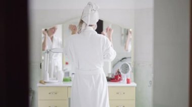 Beyaz bornoz ve saç havlusu giymiş zayıf, genç, beyaz bir kadının evdeki banyoda aynanın önünde yavaş çekimde dans edişinin arkası. Mutlu bayan sabah evde hazırlanıyor..