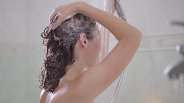 自宅でシャワーを浴びながらスローモーションでシャンプーを洗い流している若い白人女性の側面図。美しい千年紀の女性の洗濯頭と体内のバスルームで。衛生的で日常的な. — ストック動画