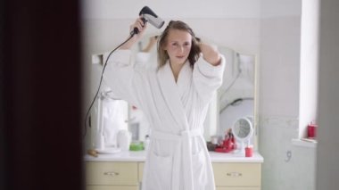 İnce, mutlu bir kadının portresi dans eden, saç kurutma makinesiyle dans eden, ağır çekimde. Beyaz bornozlu, rahat, kendine güvenen, bin yıllık beyaz bir bayanın sabah evde eğlendiğinin ön görüntüsü..