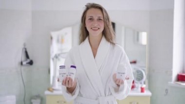 Kameraya bakıp gülümseyen, elinde vitamin takviyesi şişeleriyle gülümseyen zayıf, güzel bir kadının portresi. Beyaz tenli kadınların temel besin maddelerini tanıtırken orta açıdan çekilmiş bir fotoğrafı..