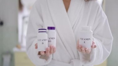 Evdeki banyoda elinde D3 E ve B vitamini şişeleri tutan tanımlanamayan ince beyaz kadın. Sağlıkla ve güzellikle ilgilenen kendine güvenen kadın yavaş çekimde içeride poz veriyor..