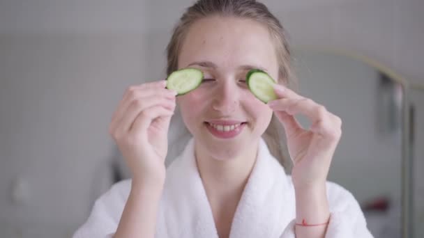 Joven hermosa alegre con sonrisa dentada divirtiéndose jugando con rodajas de pepino sobre los ojos mirando a la cámara. Retrato de alegre señora positiva delgada caucásica con máscara facial de limpieza natural. — Vídeos de Stock