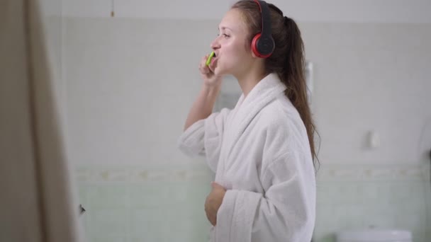 Πλευρική άποψη της νεαρής λεπτής γυναίκας με μπουρνούζι βουρτσίζει τα δόντια το πρωί στο μπάνιο στο σπίτι. Μέτριο πλάνο της όμορφης μελαχρινής καυκάσιας κυρίας με ακουστικά που απολαμβάνει το πρωί. Στοματική υγιεινή. — Αρχείο Βίντεο