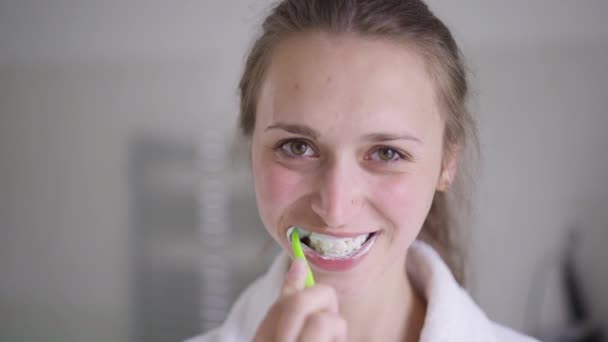 Крупный план брюнетки молодой женщины с карими глазами, чистящей зубы в замедленной съемке, улыбающейся, глядя в камеру. Портрет уверенной худой красивой белой женщины, заботящейся о стоматологическом здоровье дома. — стоковое видео