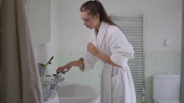 Mujer caucásica encantadora delgada en albornoz blanco que enciende el grifo y lava la cara en cámara lenta. Vista lateral de la hermosa mujer milenaria refrescante piel facial en la mañana en casa en el interior. — Vídeo de stock