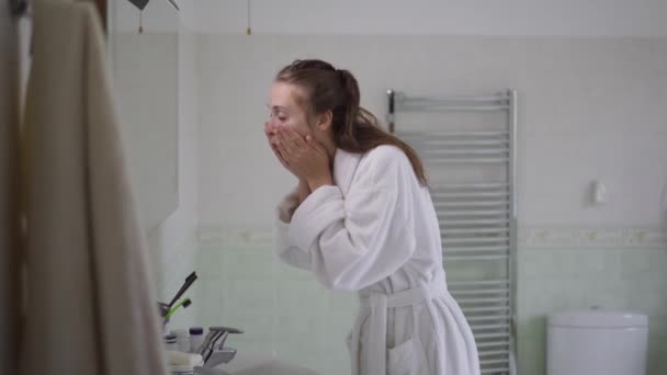 Side view portrait of young slim woman washing face with clear water in bathroom at home in the morning. Caucasian beautiful lady in white bathrobe waking up indoors cleaning facial skin. — Stock Video