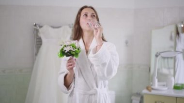 Gülümseyen beyaz bir gelinin, düğün günü şampanya içip çiçek koklamasının ön görüntüsü. Evde bornozlu, kendinden emin mutlu bir kadının portresi..