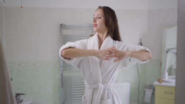 Vista lateral da mulher sorridente magro em roupão de banho branco alongando-se em pé no banheiro pela manhã. Retrato de feliz confiante bela senhora morena caucasiana acordar em casa dentro de casa. Movimento lento. — Vídeo de Stock