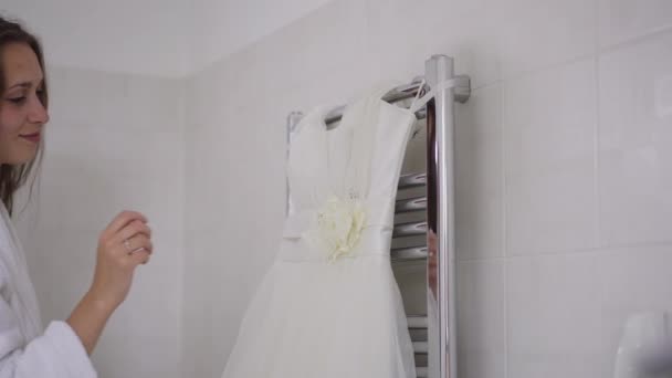 Seitenansicht des Hochzeitskleides hängen im Badezimmer mit lächelnden glücklich selbstbewusste junge Frau Eingabe berühren weiße elegante luxuriöse Kleidung. Glückliche Braut bewundert Kleid in Hochzeitsmorgen. Zeitlupe. — Stockvideo