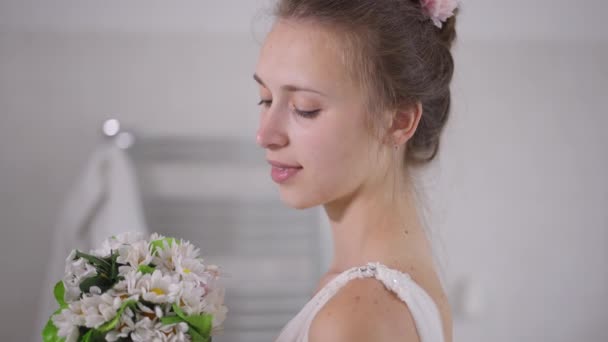 Seitenansicht Nahaufnahme Porträt der glücklichen charmanten jungen kaukasischen Braut, die weißen Brautstrauß bewundert und lächelnd in die Kamera schaut. Selbstbewusste schöne schlanke Frau posiert morgens drinnen. — Stockvideo