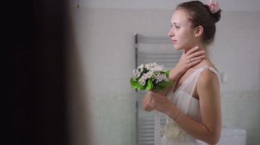 Kendine güvenen, gururlu, gelinlikli genç bir gelinin portresi. Evde banyoda buketle dikilmiş gülümsüyor. Yavaş çekimde aynada yansımasına hayran olan mutlu ince beyaz kadın..