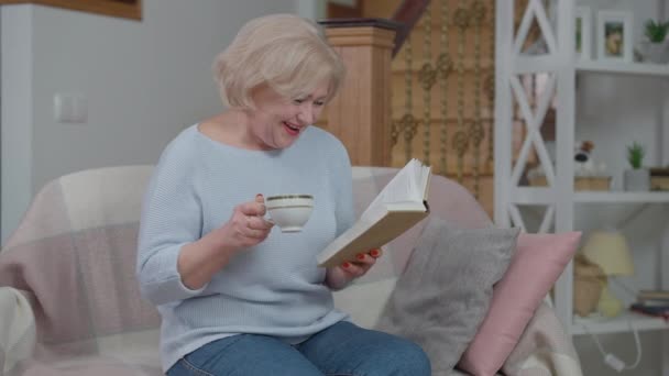 Joyful sênior bela mulher rindo leitura livro beber chá em casa na sala de estar. Retrato de aposentado caucasiano alegre se divertindo desfrutando de hobby dentro de casa. Aposentadoria e estilo de vida. — Vídeo de Stock