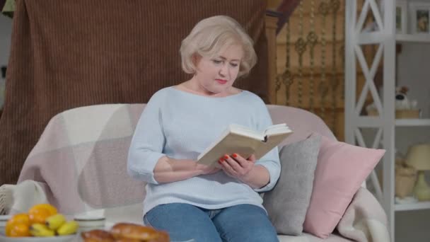 Gevşemiş yaşlı kadın kitap okurken battaniyeli, gülümseyen ve konuşan bir erkek gibi. Olgun bir anneyle ilgilenen beyaz bir çocuğun portresi evde hobi olarak eğleniyor. — Stok video