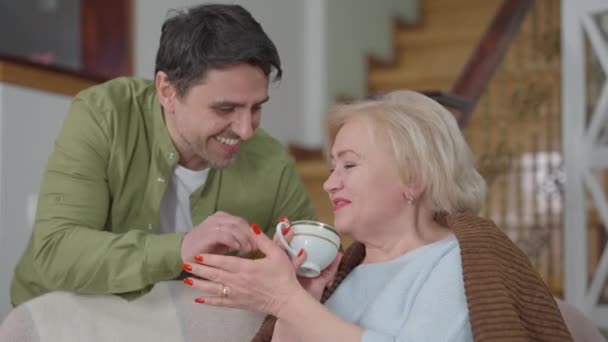 Potret wanita tua menawan yang tersenyum sambil memegang cangkir teh berbicara dengan pria dewasa yang duduk di ruang tamu. Selamat Kaukasia ibu menikmati akhir pekan dengan anak di dalam ruangan. — Stok Video