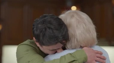 Kameraya bakıp gülümseyen, sarılan, kır saçlı, yaşlı bir annenin yakın plan portresi. Yakışıklı, neşeli, beyaz adam evde ebeveyn pozu veriyor..