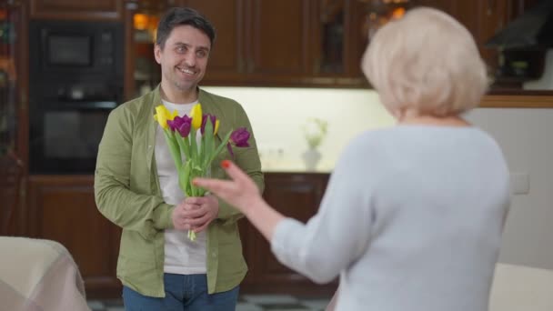 Счастливый красивый улыбающийся взрослый мужчина дарит букет желтых и фиолетовых тюльпанов пожилой женщине в помещении. Портрет кавказского сына, поздравляющего маму с днем рождения или Днем женщины. — стоковое видео