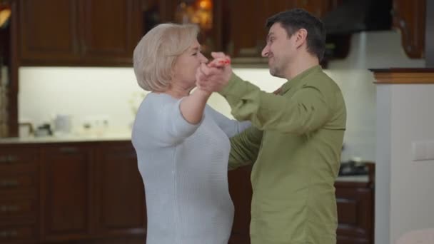 Średnie ujęcie radosnego dorosłego syna tańczącego ze starszą matką wirującą rodzicem. Portret radosnego białego mężczyzny i kobiety cieszących się wypoczynkiem w domu i zabawą. — Wideo stockowe