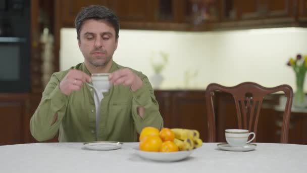 Potrait av ledsen ensam kaukasisk man dricker örtte tittar på tom stol och kopp på bordet. Upprörd deprimerad kille tillbringar helgen ensam hemma. Ensamhet och livsstil. — Stockvideo