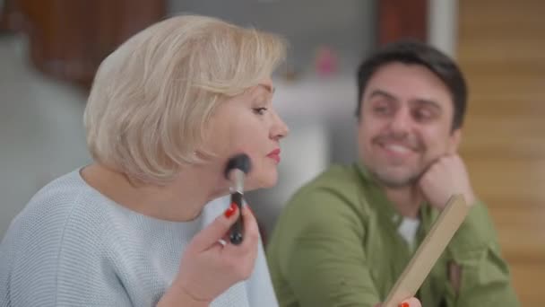 Side view portrait of positive confident senior woman applying make-up getting ready for a date with blurred smiling adult son admiring mother at background. Happy supported retiree enjoying life. — Stock Video