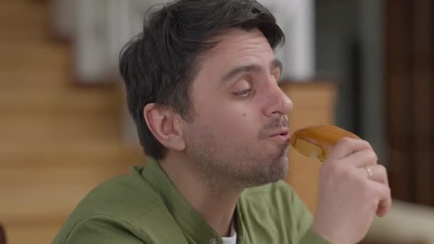 Retrato de cerca del apuesto joven caucásico disfrutando del sabor de la deliciosa pastelería casera recién horneada en el interior. Feliz chico morena relajado comiendo bollo sabroso en casa en la sala de estar. — Vídeo de stock