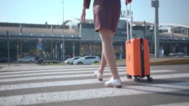Bir kadın havaalanı terminalinin önündeki yaya geçidinde yürüyor turuncu bir bavul çekiyor ve gün ışığında seyahat anını gösteriyor..